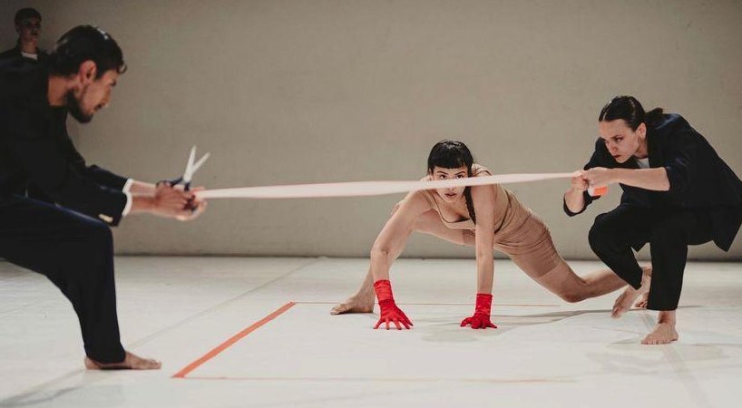 Two performers in suits holding a ribbon over a crouching dancer in red gloves
