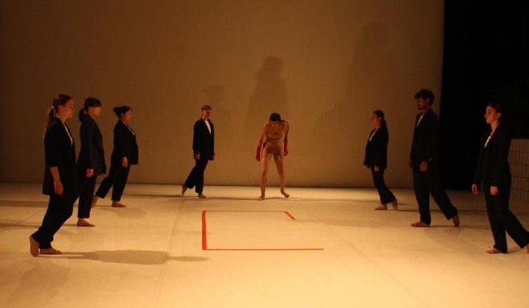 Performers in black suits standing around a lit square on stage